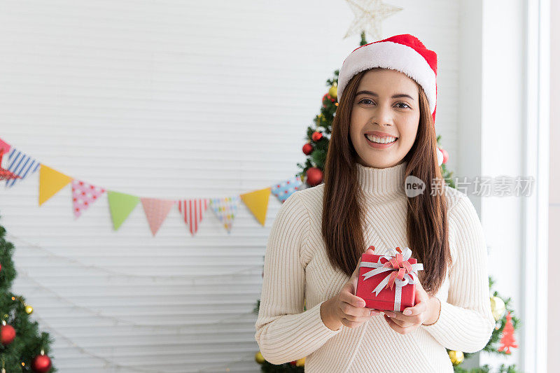 Smiling brunette woman in sweater and Santaâs hat holding small gift box and rejoices over Christmas decorative white background